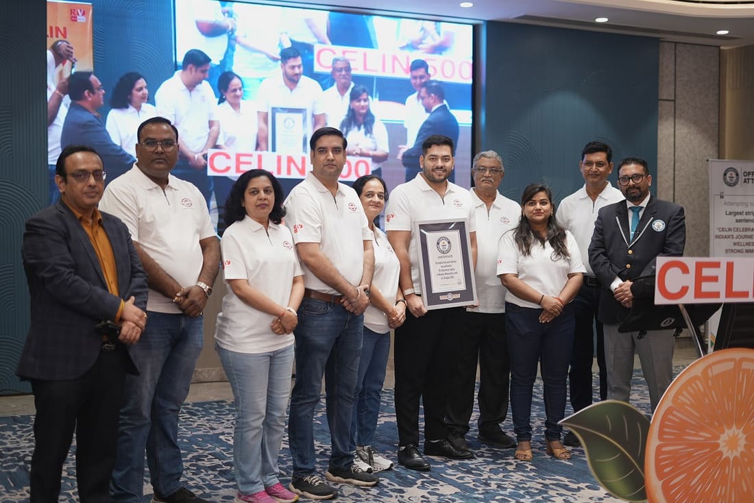Eleven members of the RV Lifesciences team standing together at the record attempt, most in white Celin polo shirts, one holding the framed Guinness World Records certificate. Behind them a screen shows the attempt in progress and a Celin 500 banner; a cut-orange display stands at the right.