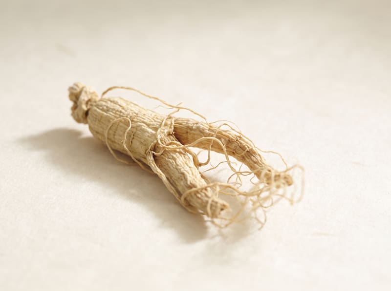 A dried ginseng root with fine fibrous tendrils on a warm cream surface.