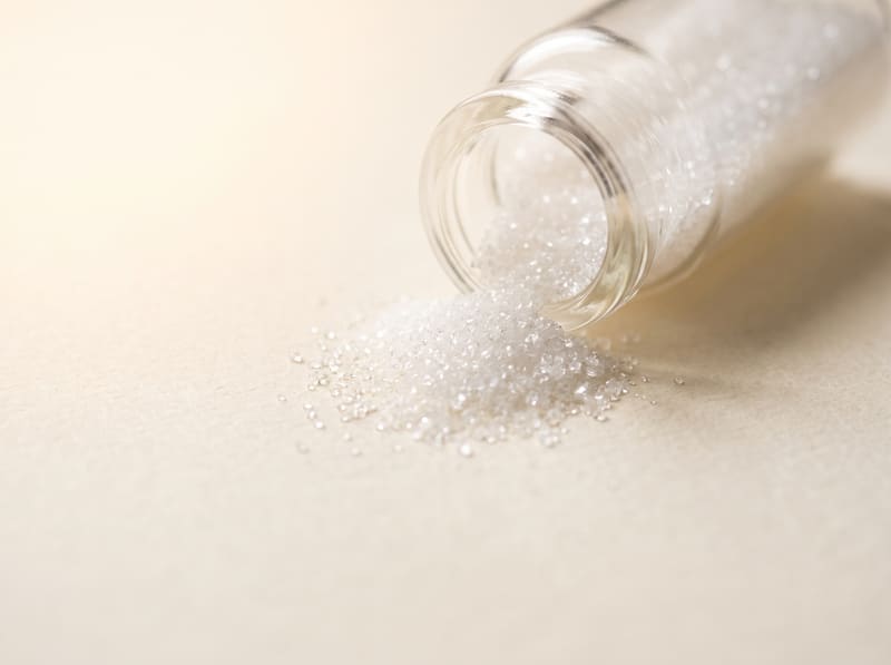 Fine white ascorbic acid crystals spilling from a small glass vial onto a warm cream surface.