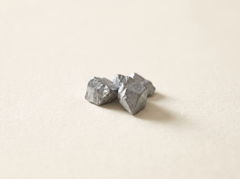 Small raw zinc metal fragments with cool grey crystalline faces, on a warm cream surface.